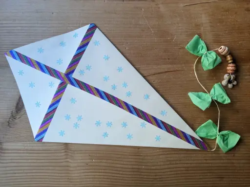 Bastelanleitung mit Bildern: Ein selbstgebastelter Drachen aus Papier mit Schneeflocken und buntem Klebeband, geschmückt mit grünen Schleifen und Holzperlen.