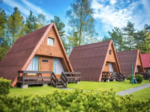 Ustronny domek nad jeziorem, jeden z kilku drewnianych domków w kształcie litery A, otoczony zielenią i drzewami.