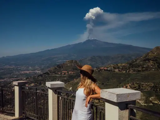 Frau mit Hut blickt auf den rauchenden sizilien vulkan. Die Landschaft ist grün und bergig.