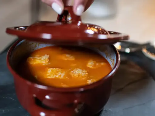 Una mano levanta la tapa de una cazuela de barro con una deliciosa comida típica de Ávila, un guiso naranja con trozos blancos.