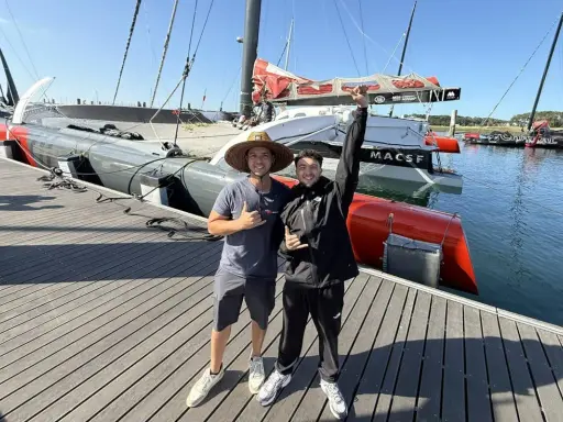 Deux jeunes hommes posent devant un bateau de course prêt pour une traversée atlantique. Le bateau est long, gris et rouge, avec des voiles partiellement visibles.