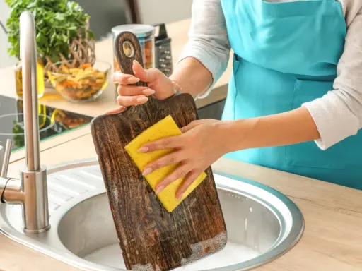 Frau reinigt ein Holz-Schneidebrett mit einem gelben Schwamm in der Spüle. Backpulver hilft, hartnäckige Flecken zu entfernen.