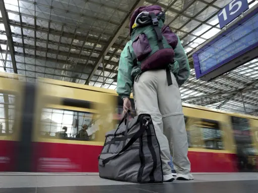 Traveler with backpack and duffel bag waits on a platform as a train speeds by.