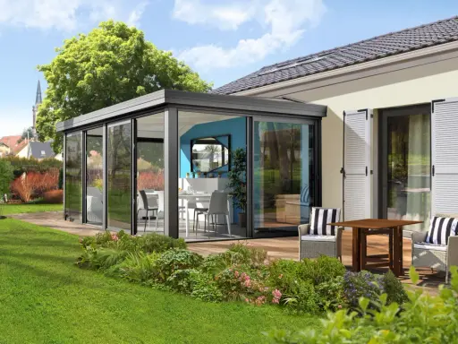 Moderne veranda sans permis de construire, avec table et chaises, offrant une vue sur un jardin verdoyant et des maisons.