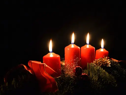 Fyra röda adventsljus brinner i mörkret, en del av en julkrans med granris och röda band. Detta är en visuell representation av vad betyder advent.