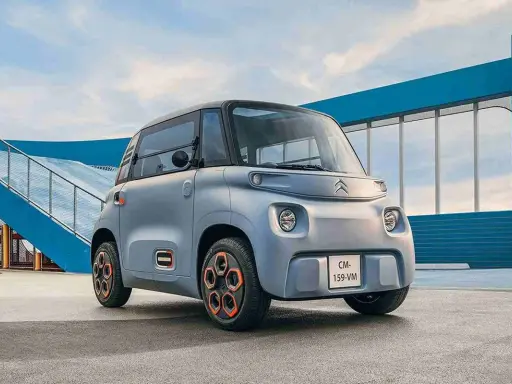 Un cuadriciclo pesado Citroën Ami gris con ruedas naranjas, aparcado frente a una estructura azul y una rampa.