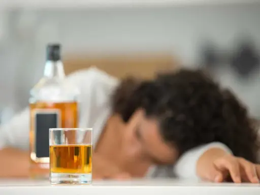 Szklanka z alkoholem i butelka na stole, osoba śpiąca w tle.
