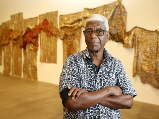 A distinguished man with white hair and glasses, arms crossed, stands before a wall adorned with shimmering, textured artworks, a testament to the enduring legacy of famous artists today.