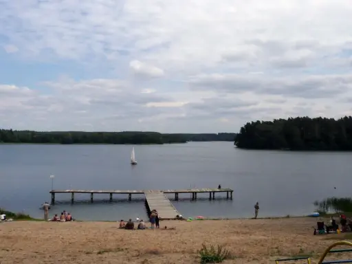Spokojny dzień na plaży nad jeziorem, żaglówka na wodzie, drewniane molo. Pojezierze Brodnickie zaprasza do wypoczynku.