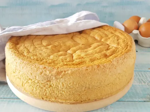 Un soffice pan di spagna dorato, coperto da un canovaccio, con uova fresche sullo sfondo.