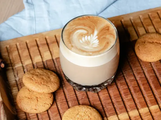 Ein Glas Flat White mit Latte Art neben Keksen auf einem Holztablett. Der Unterschied zwischen Flat White und Cappuccino ist hier deutlich sichtbar.