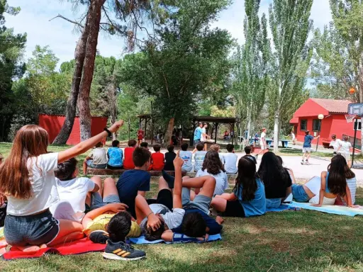 Niños disfrutando de actividades al aire libre en campamentos de verano aula joven. Un grupo observa un partido de baloncesto mientras otros descansan en el césped.