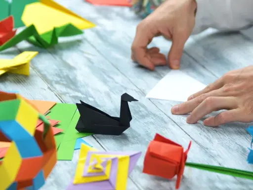 Ręce składają papier, tworząc origami: czarnego łabędzia, czerwoną różę i kolorowe figury. Pokazuje, co można zrobić z papieru.