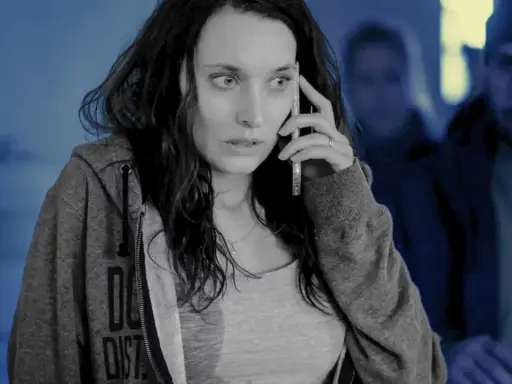 Przerażona kobieta rozmawia przez telefon, podczas gdy w tle widać zamazane postacie. Scena jak ze skandynawskich seriali kryminalnych.