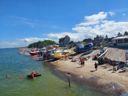 Słoneczny dzień na plaży, kolorowe łodzie na piasku, tłumy ludzi szukają idealnych miejsc na wakacje.