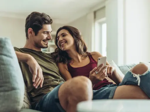 Pareja sonriente en el sofá, compartiendo un momento íntimo. Ella le muestra algo en su móvil, una forma de como expresar sentimientos.