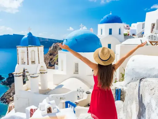 Santorini, Grecja, gdzie warto pojechać. Kobieta w czerwonej sukience i kapeluszu cieszy się widokiem białych domów i niebieskich kopuł nad morzem.