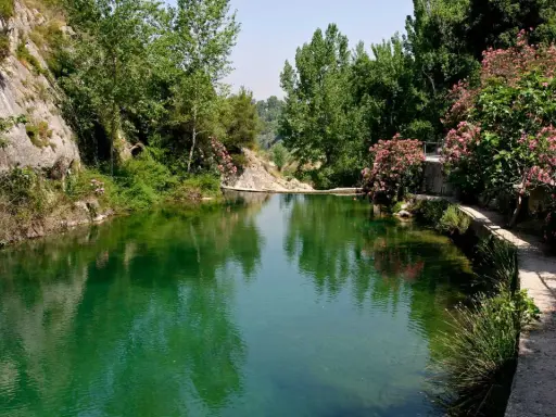 Un remanso de agua verde rodeado de rocas y vegetación exuberante. Un camino serpentea junto al agua, ideal para explorar qué ver en Ontinyent.