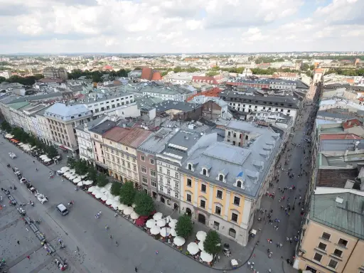 Widok z góry na Rynek Główny w Krakowie. Tłumy ludzi spacerują, a dorożki czekają na turystów. W tle widać zabytkowe kamienice i rozległą panoramę miasta.