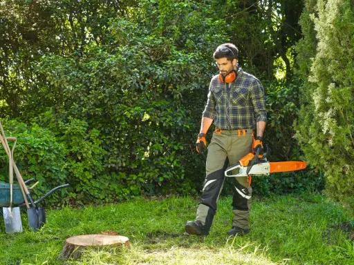 Hombre con motosierra se acerca a un tocon de arbol en un jardín. Herramientas y carretilla a la izquierda.