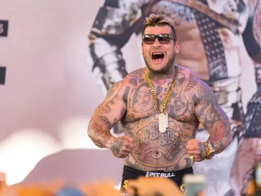 Popek kiedys, wytatuowany mężczyzna w okularach przeciwsłonecznych, z złotym łańcuchem i wisiorem, napina mięśnie.