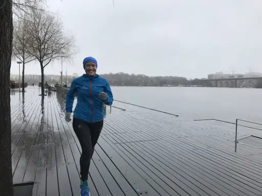 Glad kvinna med blå jacka och mössa springer i regn längs en träbrygga vid en sjö.