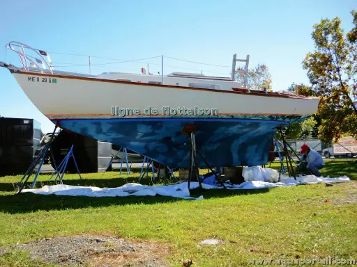 Un bateau est sur des chandelles, prêt pour une peinture. La ligne de flottaison est visible sur la coque bleue. Un homme travaille sur le bateau.