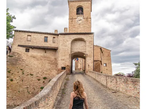 Passeggiata verso il borgo di Montegridolfo cosa vedere: un antico portale in mattoni con torre dell'orologio e un ponte acciottolato.