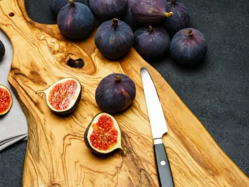 Kilka dojrzałych, ciemnofioletowych fig, niektóre przekrojone na pół, leżą na drewnianej desce obok noża.