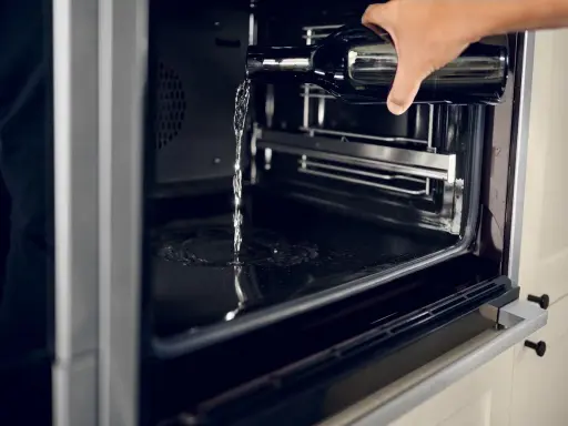 Wasser wird aus einer Flasche in einen modernen **Hydrolyse Backofen** gegossen, um die Reinigung zu erleichtern.