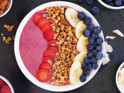 En färgglad acai bowl med jordgubbar, banan, blåbär och granola. Serverad med mandlar och kokosflingor vid sidan.
