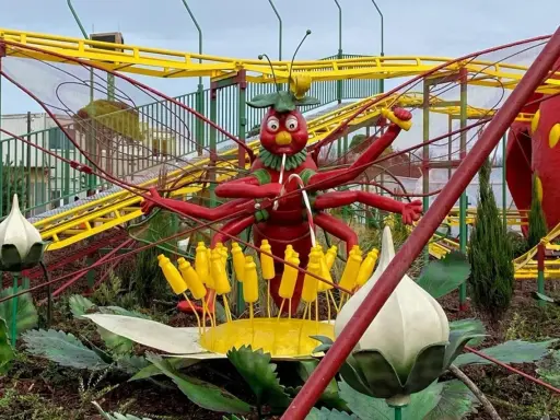 Eine rote Insektenfigur mit vielen Armen steht vor einer gelben Achterbahn.