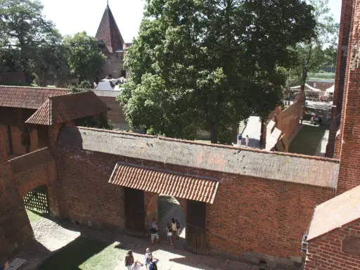 Wielki zamek w Malborku, ceglane mury, dziedziniec z drzewami i ludźmi spacerującymi pośród zabytkowej architektury.