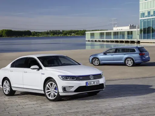 Dos coches Volkswagen, uno blanco sedán y otro azul familiar, estacionados frente a un edificio moderno junto a un lago. ¿Cuál es el permiso de coche?