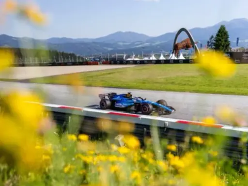 Un coche de F1 azul compite en un circuito soleado, con flores amarillas en primer plano. ¡Un gran resultado f1 hoy!
