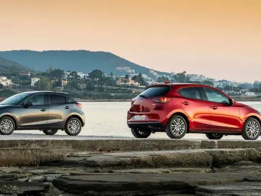 Dos Mazda, uno gris y otro rojo, junto al mar. La fiabilidad Mazda se refleja en su diseño y presencia.