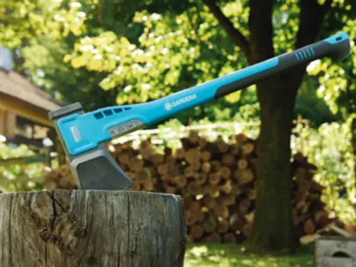 Une hachette bleue et grise est plantée dans un billot, prête à couper du bois. Une pile de bois est visible en arrière-plan.