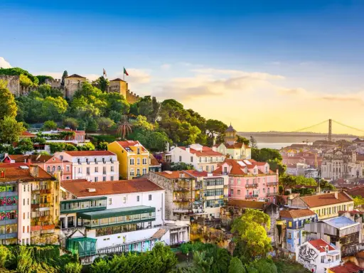 Bunte Häuser und eine Burg auf einem Hügel in Lissabon, der Hauptstadt Portugals. Im Hintergrund die Hängebrücke über den Tejo.