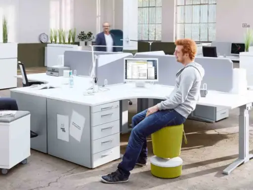 Un espace de travail open space moderne avec des bureaux modulables, des plantes vertes et des sièges colorés. Deux personnes discutent, une troisième travaille sur son ordinateur.