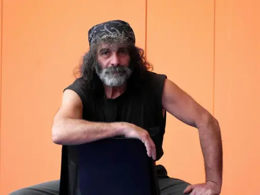 Mauro Corona, con bandana e barba grigia, seduto su una sedia. Immagina i suoi libri in ordine cronologico.