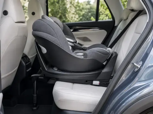 Silla de coche para bebé, instalada a favor de la marcha, con base giratoria y tapicería gris sobre asientos de cuero blanco en el interior de un vehículo.