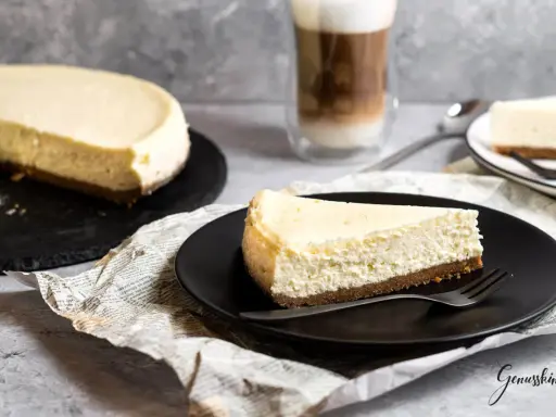 Ein Stück New York Cheesecake auf schwarzer Platte, daneben ein ganzer Kuchen und ein Latte Macchiato.