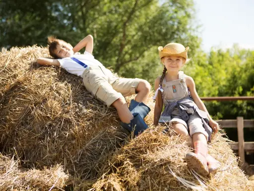 Dzieci odpoczywają na sianie podczas pobytu na agroturystyce ze zwierzętami. Chłopiec leży z rękami za głową, a dziewczynka siedzi w słomkowym kapeluszu.