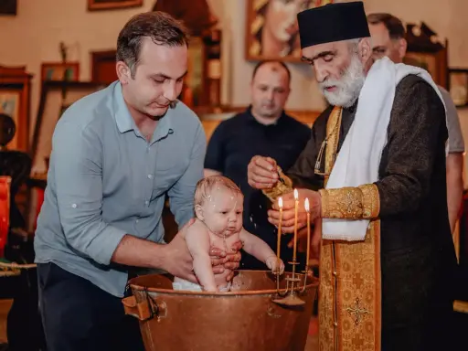 Chrzest dziecka w kościele. Ojciec trzyma niemowlę w miedzianej chrzcielnicy, a ksiądz polewa je wodą. To ważne chrzciny.