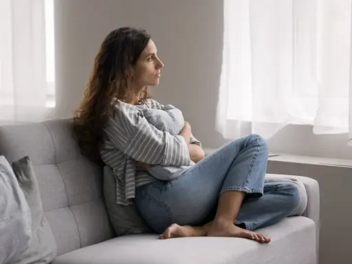 Kobieta siedzi na kanapie, przytulając poduszkę. Zamyślona patrzy przez okno. To pokazuje, po co światu psychoterapia – by odnaleźć spokój.