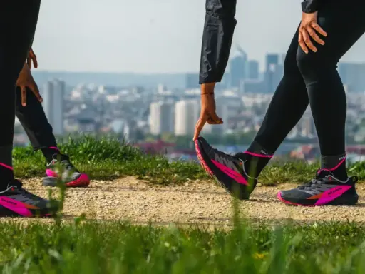 Zwei Läuferinnen dehnen sich auf einem Feldweg mit Blick auf eine Stadt. Sie tragen schwarze Laufhosen und auffällige pink-schwarze Laufschuhe. Ein Zeichen für den Wiedereinstieg ins joggen nach Geburt.