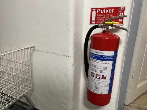Röd brandsläckare hänger på en vit vägg, redo att användas om brandlarm ljuder. Bredvid en vit metallkorg.