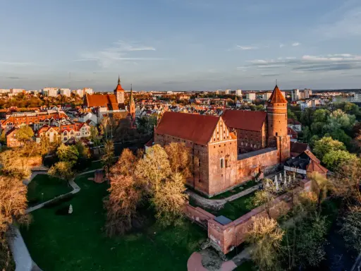 Zachód słońca nad zamkami krzyżackimi na Mazurach. Potężne ceglane mury i wieże w blasku złota, otoczone zielenią i miastem.