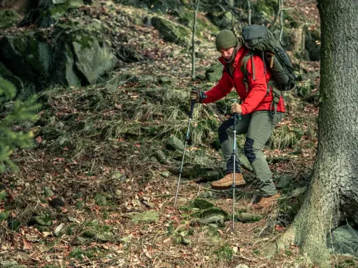Mężczyzna w czerwonej kurtce i zielonej czapce z plecakiem wspina się po kamienistym, leśnym szlaku, używając kijków. To jest właśnie trekking!