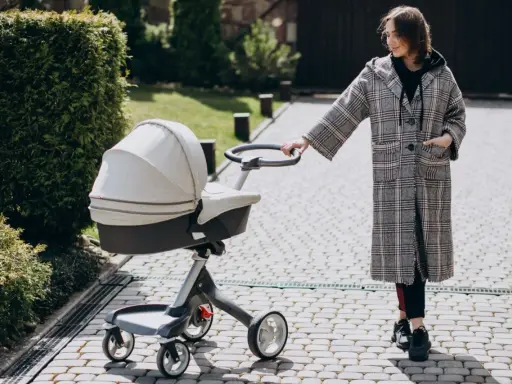Kobieta w płaszczu prowadzi spacerówkę kubelkową po brukowanej ścieżce wśród zieleni.
